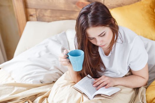 A young woman enjoys a relaxing morning in bed, reading a book with a coffee mug in hand.