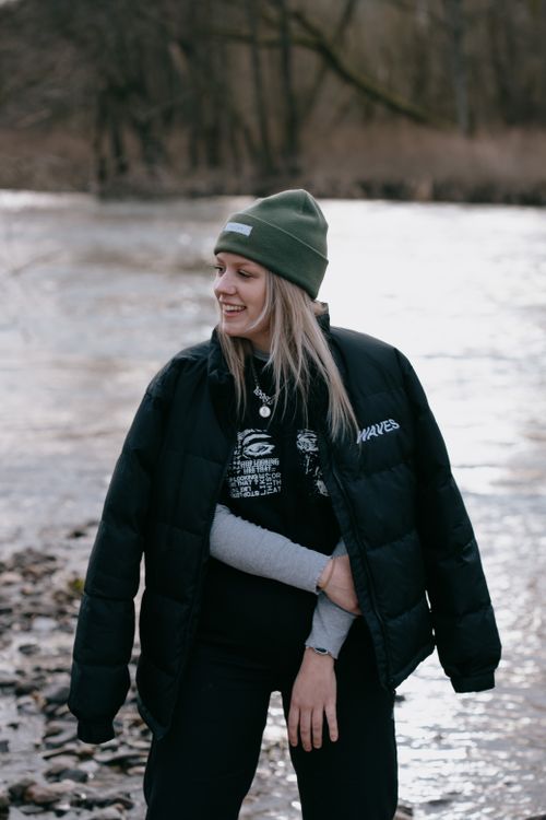 Free A woman in a beanie and puffer coat smiles by a riverside during winter. Stock Photo