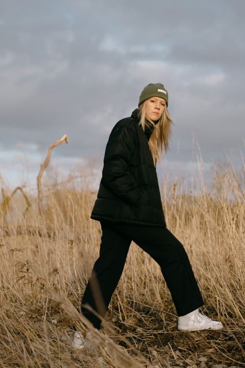 Free Woman in black jacket and green beanie walking in a Belgian field under a cloudy sky. Stock Photo