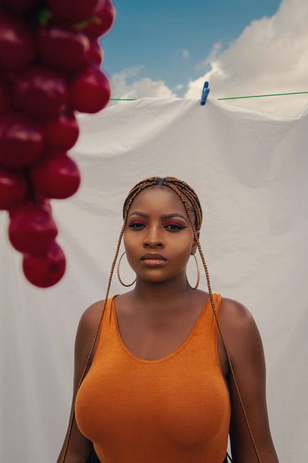 Confident young black lady wearing orange swimsuit with earrings near purple bunch of grapes with white sheet on clothesline with pin under blue cloudy sky and looking at camera