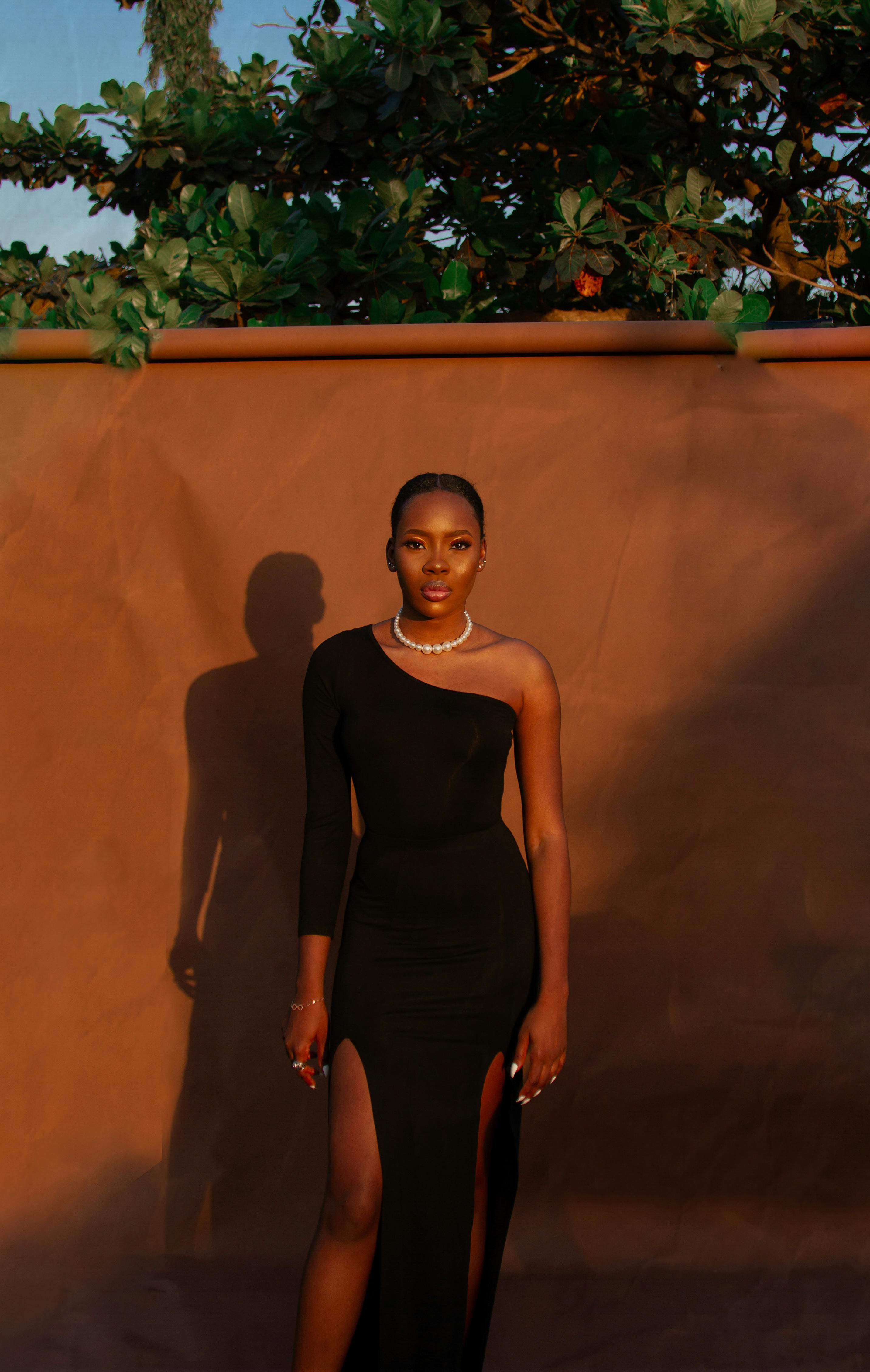 Woman in a chic black dress stands outdoors under the sun, exuding elegance and confidence.