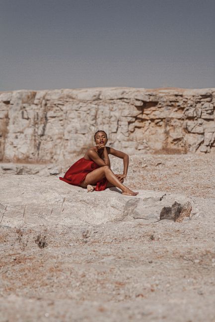Full body of young black lady in red cloths sitting on rock near sandy terrain and uneven stone hill in sunny summer day under blue cloudless sky with closed eyes