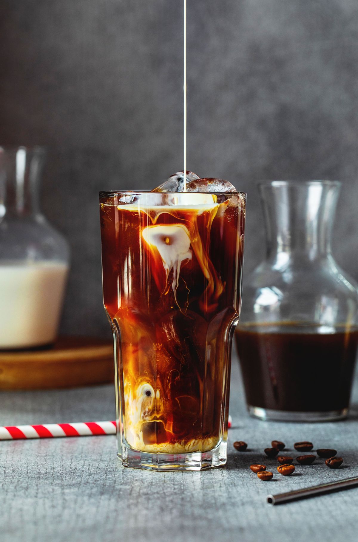 Glass of iced cold brew coffee with cream pouring in, carafes of coffee and milk behind