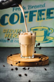 Glass with fresh coffee with milk and ice cubes placed on tray on table with coffee beans near cafe signboard