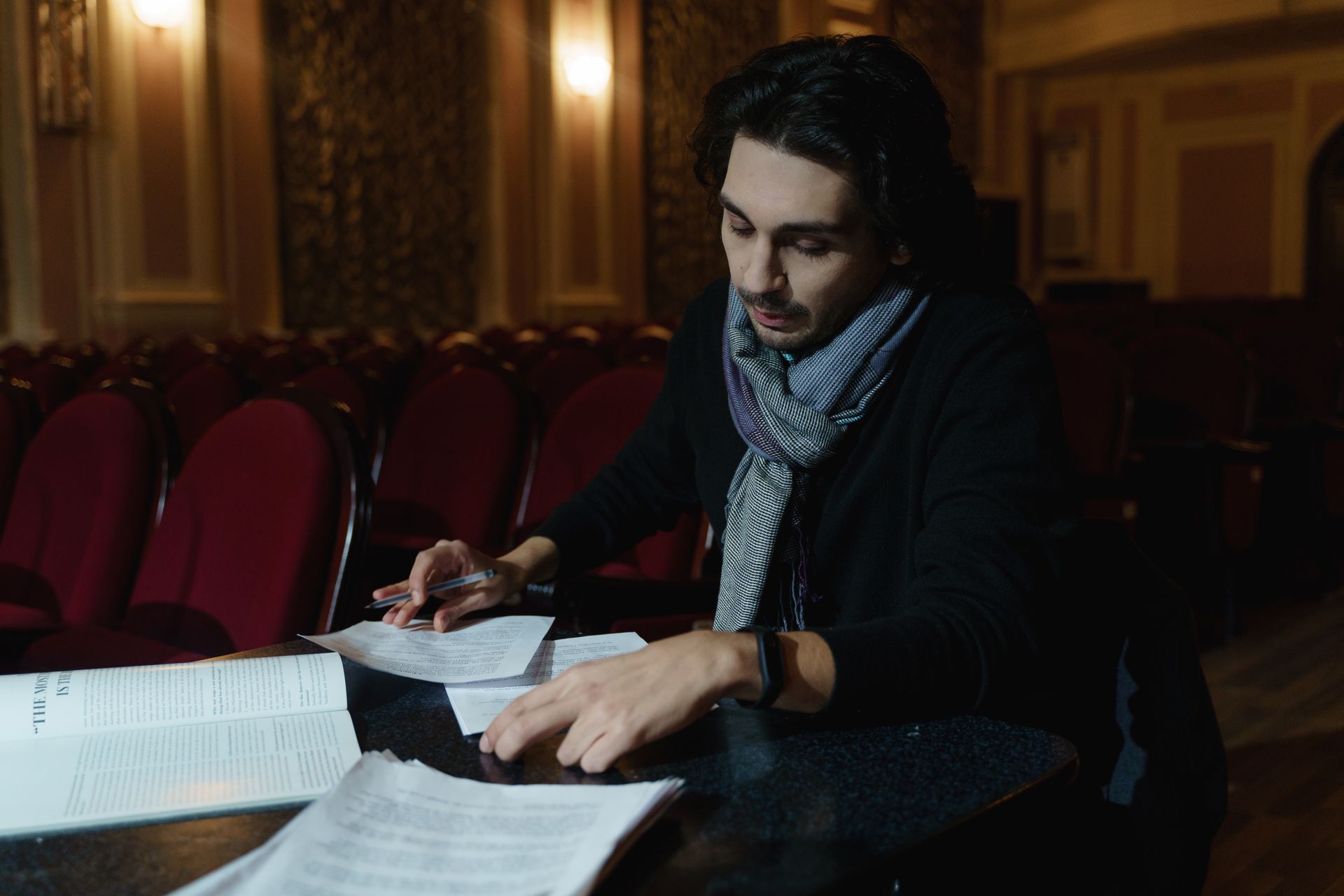 A theater director is deeply engaged in reviewing scripts in a dimly lit auditorium.