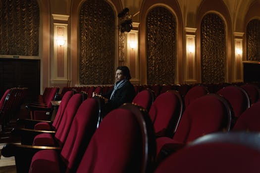 A man sits alone in an opulent theater, surrounded by red chairs and ornate decor.