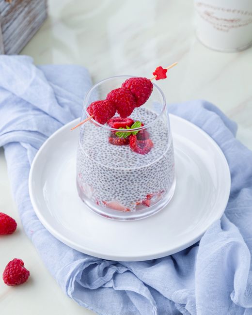 A purple-pink fruit smoothie in a tall glass topped with a sprinkle of chia seeds and a few fresh raspberries, with a reusable straw resting against the rim.