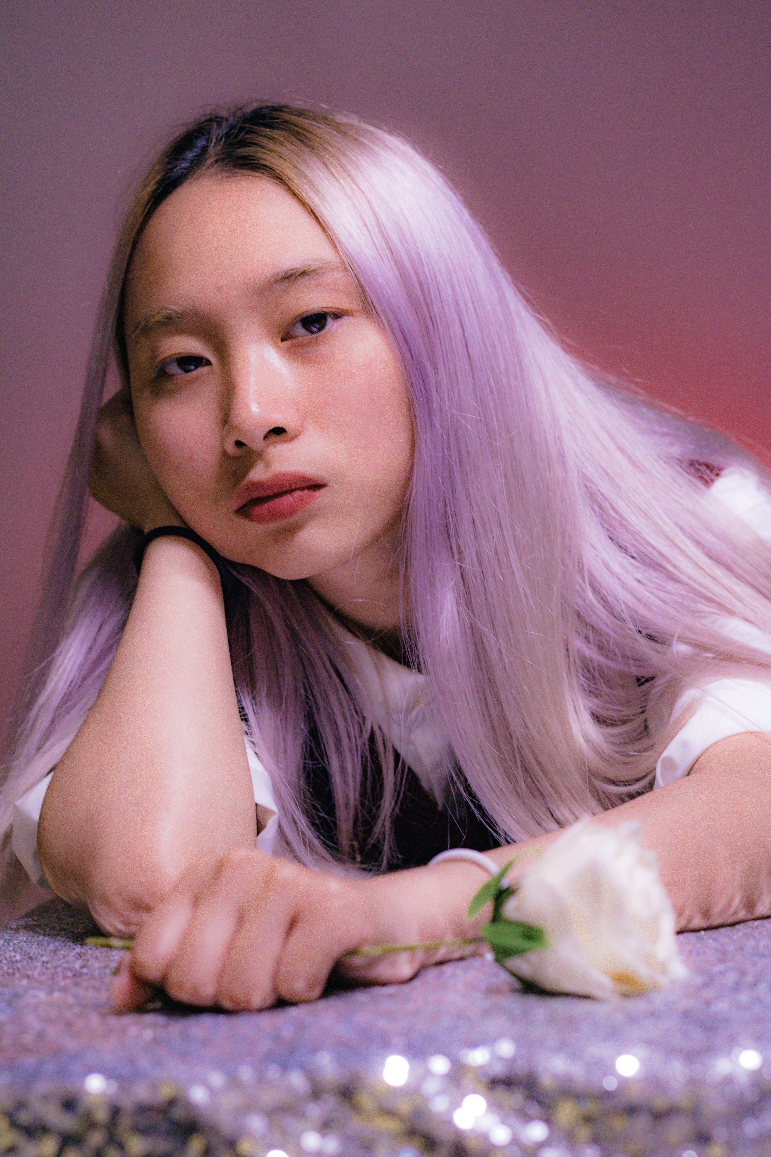 Close-up portrait of woman with lavender hair resting on sequined fabric, holding white rose.