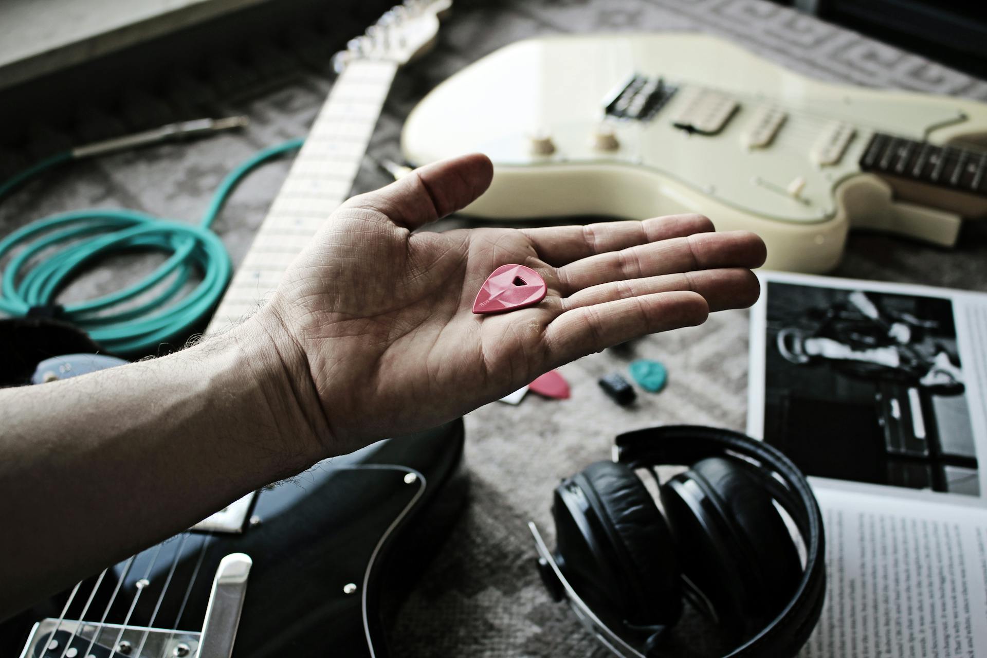 Close-up of a hand holding a guitar pick with electric guitars, cables, and headphones in the background.