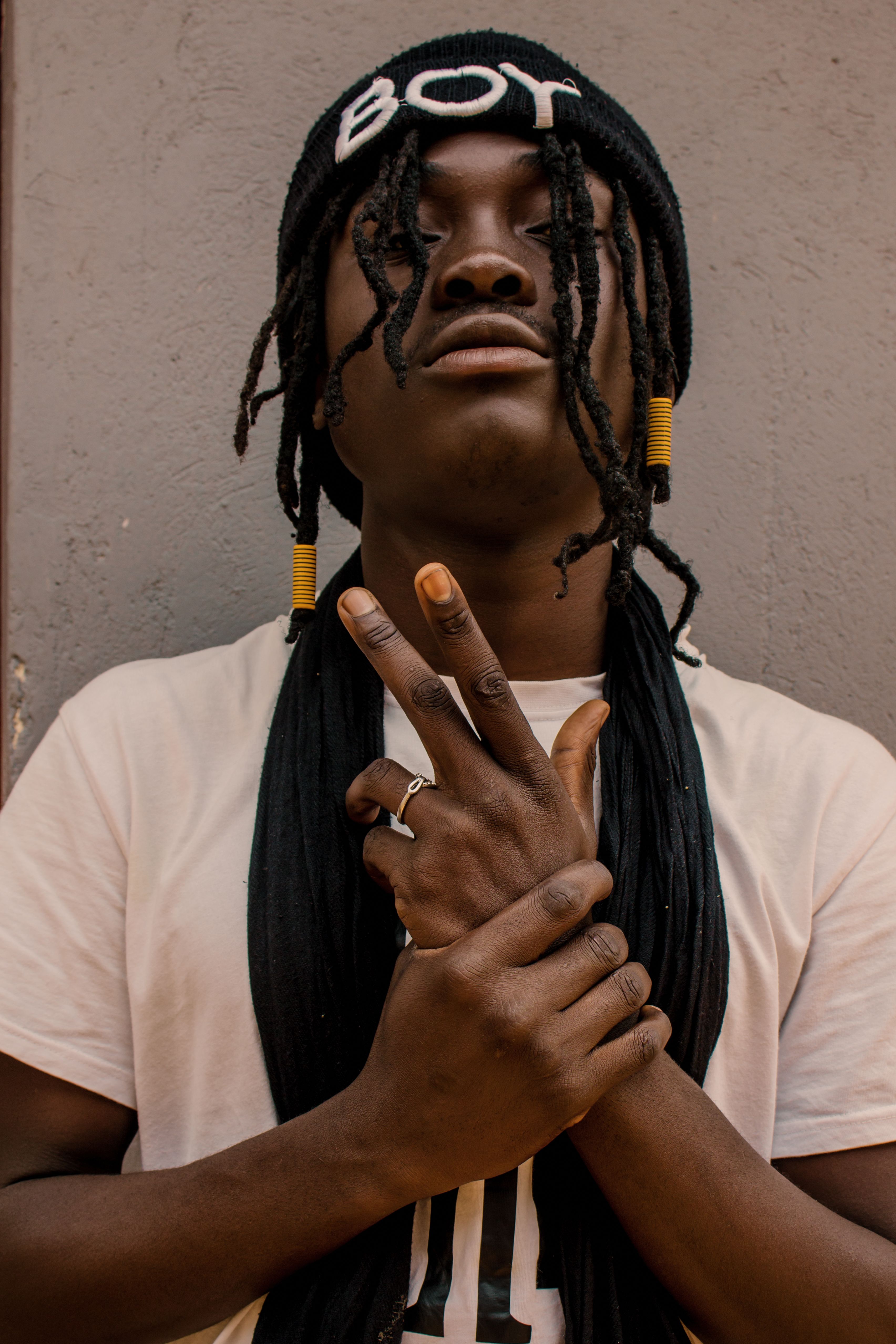 Confident young black man in t shirt and hat with scarf and dreadlocks making gun gesture with hand near stone wall with closed eyes in daytime