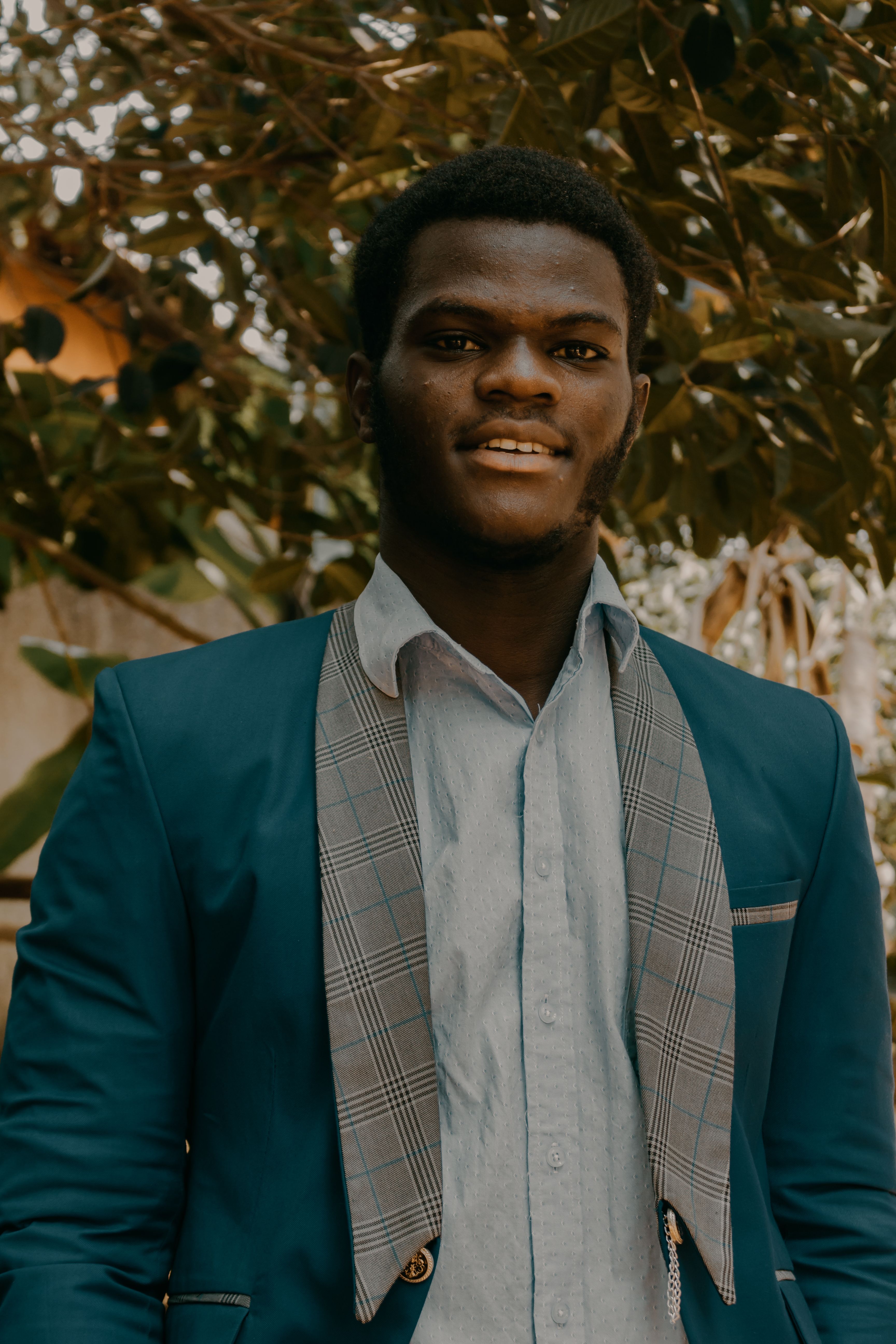 Positive young African American man in shirt and jacket looking at camera near trees and plants with green leaves in daytime in countryside