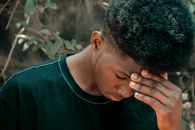 Sad young African American man in casual outfit with hand on head and closed eyes near trees and plants with green foliage in countryside in daylight