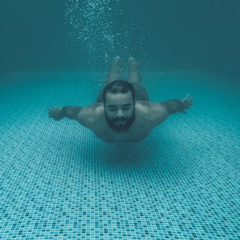 A bearded man enjoys swimming underwater in a turquoise pool, captured with a mobile camera.