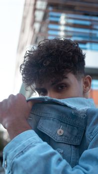 Portrait of a young man with curly hair outdoors, wearing a denim jacket, urban background.