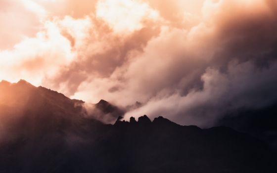 Silhouette of mountains in Austria beneath dramatic clouds and warm orange sunset.