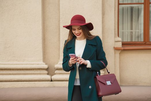 Fashionable Woman in a Bottle Green Coat and Burgundy Hat Smiling and Using Phone 