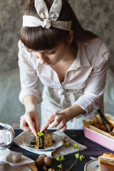 A woman decorates a cake slice indoors with fresh greenery on a wooden surface.