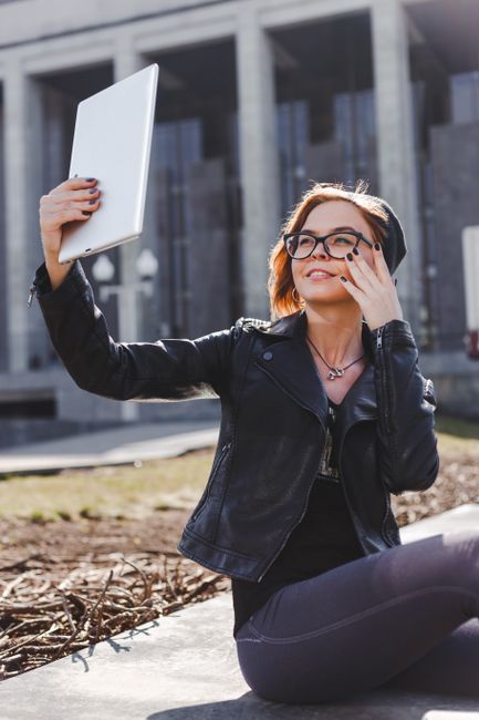 Stylish woman wearing a leather jacket and glasses takes a selfie outdoors on a sunny day.