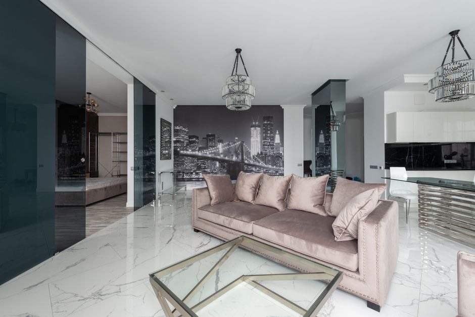 Interior of light modern flat with sofa with pillows near glass table under chandelier next to chair and open kitchen with cupboards near glass doors