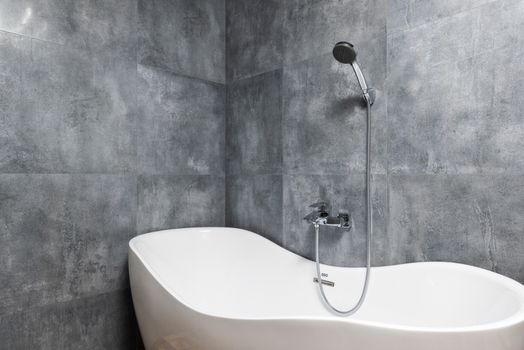 Interior of light modern bathroom with white bath under shower on gray tile wall