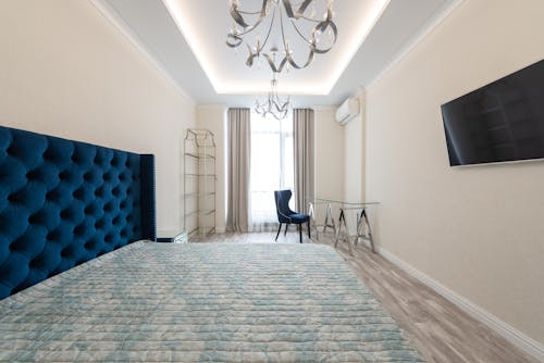 Interior of light bedroom with bed in front of TV on wall next to chair with glass table near window with curtains under chandeliers