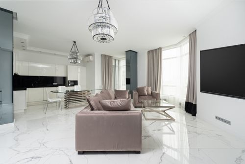 Free Interior of modern light studio with couch and armchair with pillows near glass table and chairs under chandelier next to open kitchen with white cabinets and windows with curtains Stock Photo