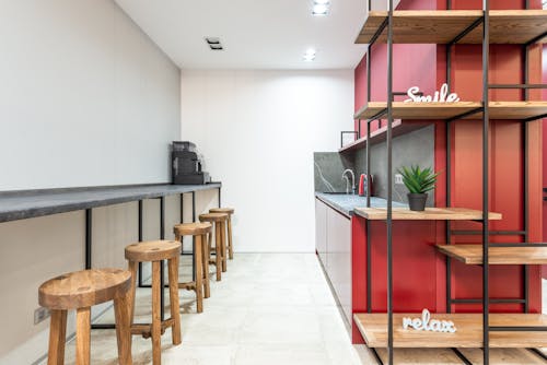 Interior of modern workplace with counter and stools next to open kitchen with cabinets near wooden shelves with potted plant and words relax and smile