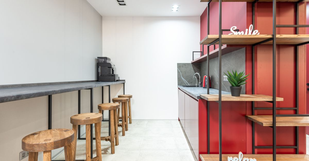 Interior of modern workplace with counter and stools next to open kitchen with cabinets near wooden shelves with potted plant and words relax and smile
