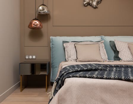 Interior of light bedroom with pillows on bed next to table under lamps hanging from ceiling