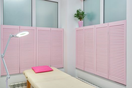 Interior of contemporary spa salon with couch under bright light of lamp with pink shutters on windows