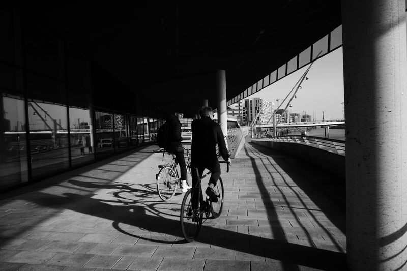 Two cyclists ride under a bridge in Hamburg, casting shadows on the pavement in a grayscale urban scene.