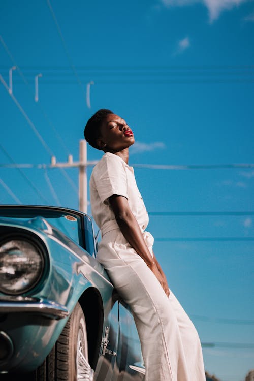 Side view of young African American female in stylish white outfit with closed eyes and bright red lips leaning on vintage car on blurred background of clear blue sky