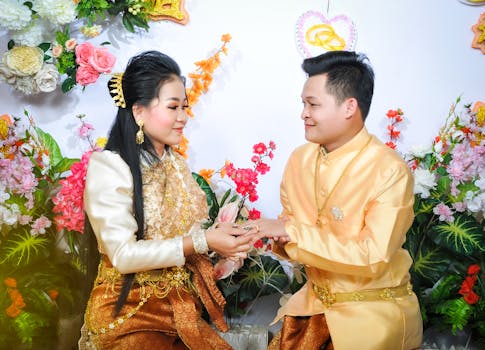 A Cambodian couple in traditional wedding attire exchanging rings amidst colorful floral decorations.