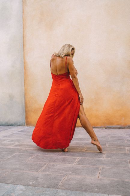 A graceful woman dancing barefoot in a flowing red dress against a textured wall.