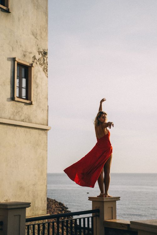 Free Elegant woman in a red dress gracefully dancing on a seaside balcony with ocean view. Stock Photo