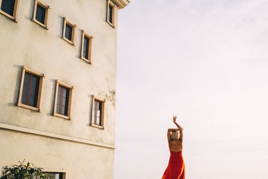 A woman in a red dress stands outdoors beside a tall building during sunset.