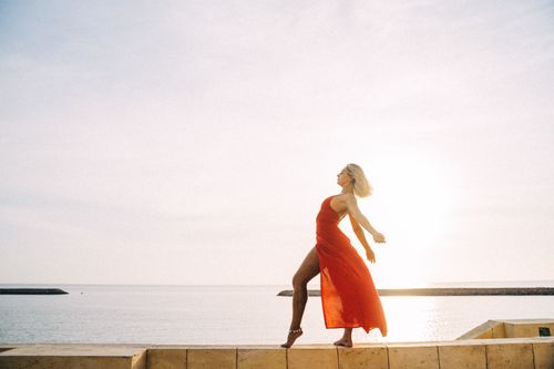 Free A woman gracefully dances in a red dress by the sea under a clear sky, showcasing freedom and elegance. Stock Photo
