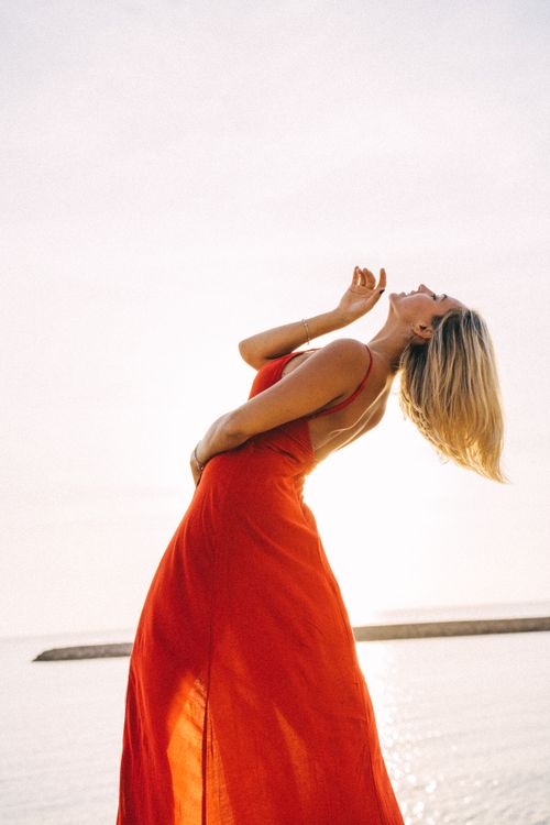Free Graceful woman in a flowing red dress captured mid-dance by the sea. Stock Photo