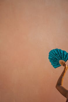 A partially visible woman elegantly holds a blue hand fan against a textured wall.
