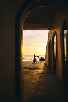 A dancer in a red dress silhouetted by a sunset on a beach, framed by an archway.