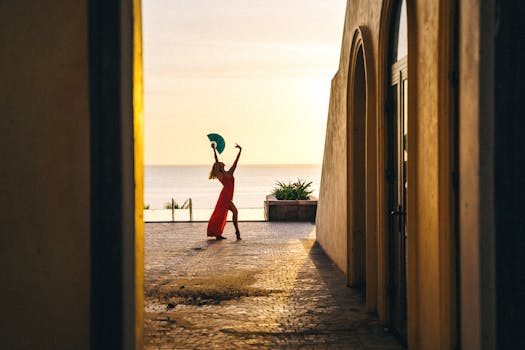 Elegant silhouette of a dancer in a red dress by the sea at sunset, framed by Mediterranean architecture.