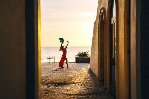 Free Elegant silhouette of a dancer in a red dress by the sea at sunset, framed by Mediterranean architecture. Stock Photo