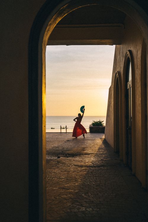Free A silhouette of a woman elegantly dancing with raised hands by the beach at sunset, framed by an archway. Stock Photo