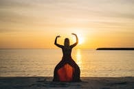 Back view of female with raised hands doing yoga standing on sandy beach with face towards calm sea and sunset in summertime