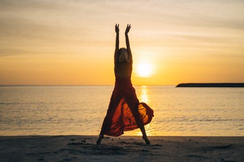 Free A woman stands on the beach at sunset, arms raised, creating a peaceful silhouette against the ocean. Stock Photo