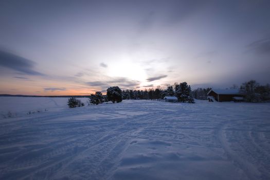 A serene winter sunset over a snow-covered rural countryside with trees and a house.