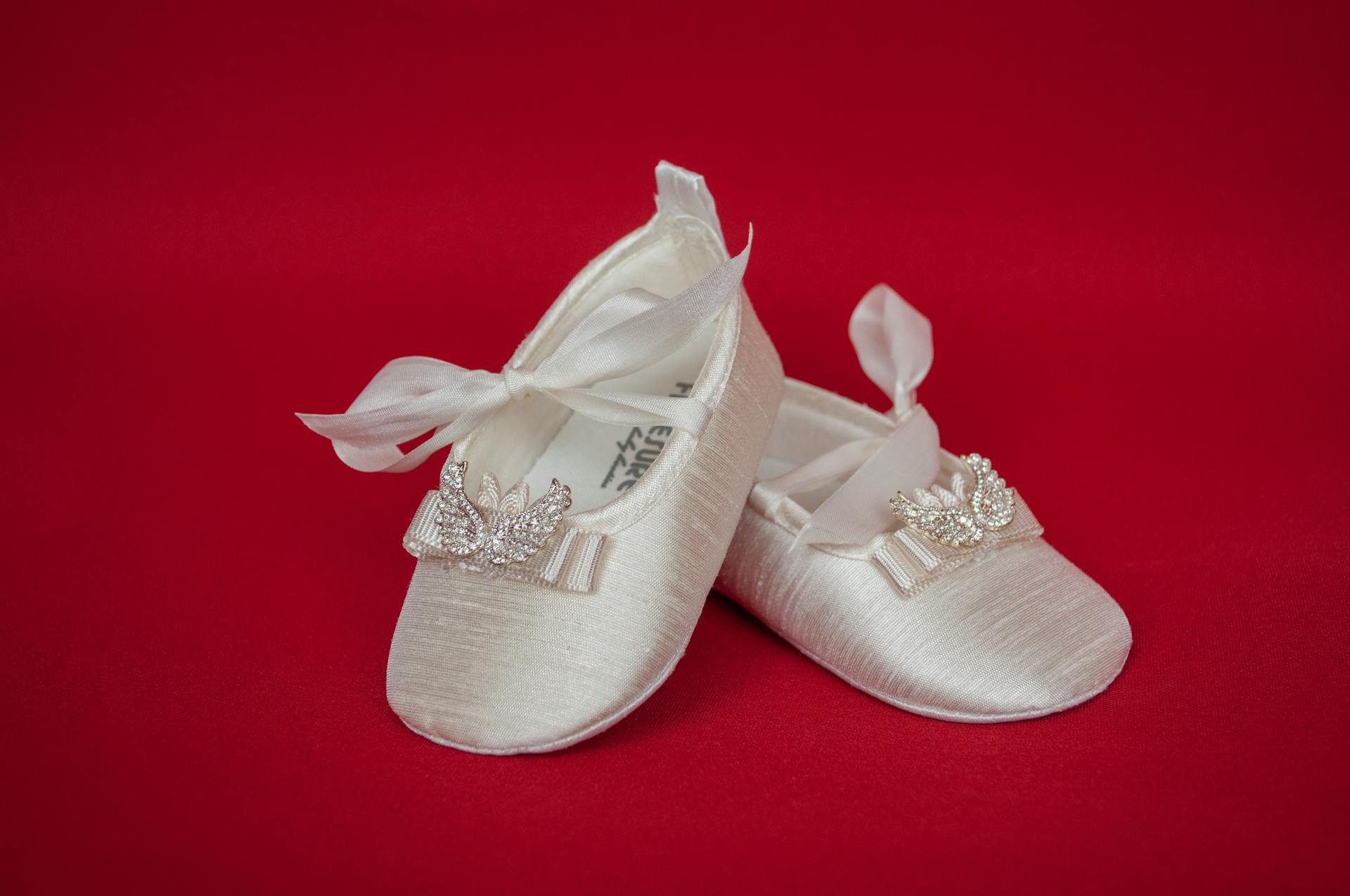 Close-up of elegant satin baby shoes with bow and embellishments on a red background.