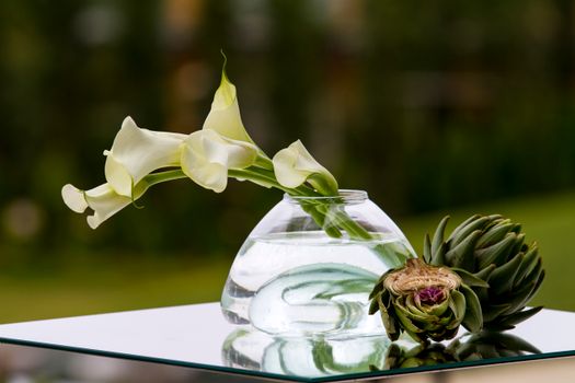 A minimalist still life of elegant calla lilies in a glass vase with an artichoke on a reflective surface.