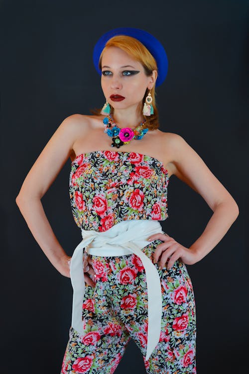 Self assured young lady with red lips in stylish outfit and beret standing against black background with hands on waist and looking away