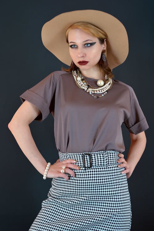 Self esteem young female model in stylish outfit and hat with bright makeup standing against black background with hands on waist and looking away